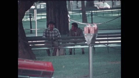 1970s Men sitting on bench in park. Kids playing with fire hydrant in street