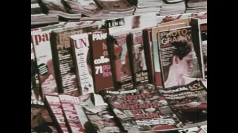 1970s: Sidewalk.  People walk down street.  Man stands at table.  Magazines.  Stockbeeldmateriaal 292808040