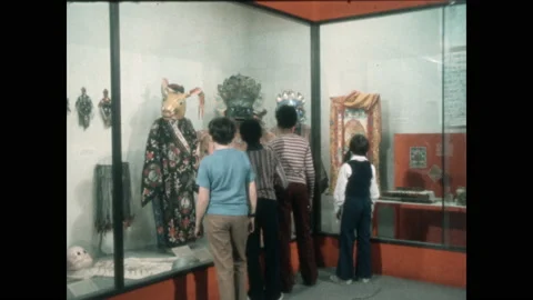 1970s: Teens look in display case at museum. Carved masks on embroidered robes. Vidéo 154697914