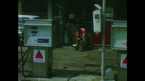 1970s: Young man sits on crate at gas station. Car pulls up to pump at station Stock Footage