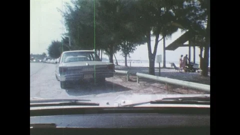 1980s: car parallel parks on beach road. man in striped shirt gathers towel, Stock Footage 77846691