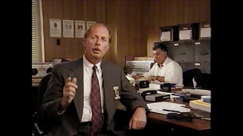 1990s: man with badge sits, talks and gestures in office while man behind him Stock Footage 128194345