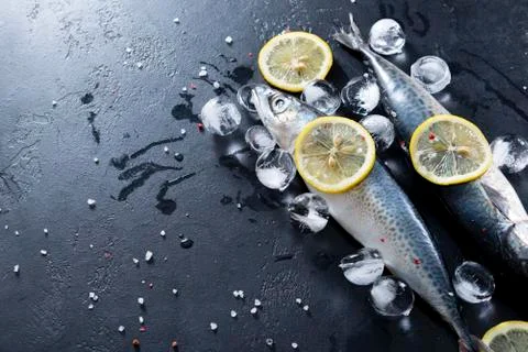 2 fresh raw fish mackerel, lemon slices, ice cubes, spices, salt on a black b Stock Photos