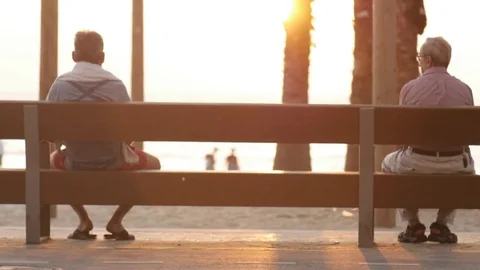 2 old men sitting alone, separate on a bench at the beach watching sunset Stock Footage 75397617