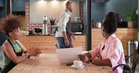 2 women sitting at table in cafe, woman reading docs aloud, aproned man coming Stock Footage 338168128