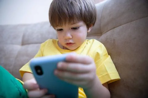 2 year old boy is sitting on the couch and playing on the phone. Little cute boy Stock Photos