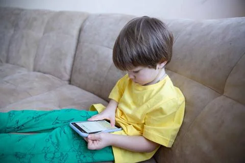 2 year old boy is sitting on the couch and playing on the phone. Little cute boy 写真素材