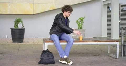 20s man sitting on bench by backpack, reaching for orange drink, eating sandwich Stock Footage 345062595