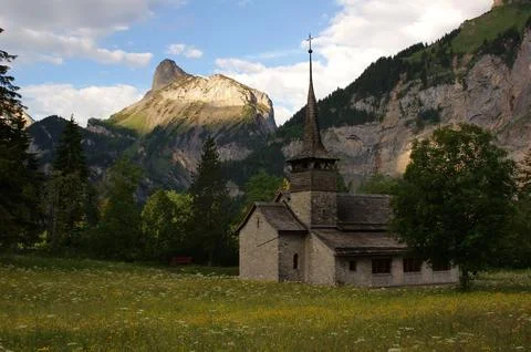 A scenic view of an old church located in a field surrounded by mountains in Kandersteg, Switzerland xkwx ancient, architecture, background, building, church, clouds, field, foliage, grass, green, leaves, meadow, mountains, old, religion, scenic, sky, trees