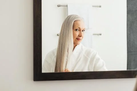 Portrait of an aged woman with long grey hair. Female looking in the mirror admires her hair. xkwx admires, adult, aged, bathrobe, care, caucasian, grey, grey hair, indoors, lifestyle, long hair, looking, mature, mirror, morning, one, people, person, portrait, real, reflection, routine, senior, woman
