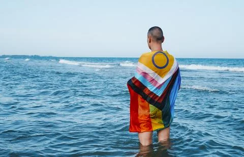a wet person in the sea wrapped in an intersex-inclusive progress pride flag standing in the water, as seen from behind xkwx intersex inclusive progress gay pride flag person man symbol rainbow LGBT LGBTI LGBTQ LGBTIQ homosexual homo homosexuality lesbian transgender transgenderism trans transsexual transsexualism intersexual bisexual bisexuality gender identity orientation queer genderqueer non-binary non binary enby diversity activism activist rights same sex sexual sexuality freedom wet sea ocean outdoors summer horizontal