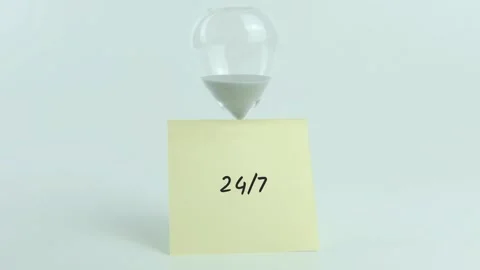 24/7 written on yellow sticky note placed on hourglass on white background Stock Footage 313459618