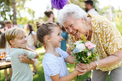 Garden birthday party for senior lady. Beautiful senior birthday woman receiving flowers from granddaughter and grandson. xkwx birthday woman senior flowers gifts garden birthday party beautiful woman lady receiving gift bouquet friends family grandmother grandchildren celebration joy happiness presents outdoors event festivity gathering special occasion surprise smiles enjoyment togetherness love affection decorations cake memories balloons laughter congratulations wishes entertainment bonding delight cheerful atmosphere friendship gratitude copy space gray hair pearls