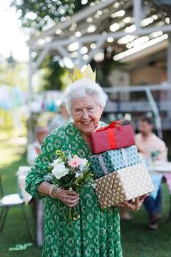 Garden birthday party for senior lady. Beautiful senior birthday woman with paper crown on head and gifts in hands. xkwx birthday woman senior crown paper flowers gifts garden birthday party beautiful woman lady receiving gift smile friends family grandmother celebration joy happiness presents outdoors event festivity gathering special occasion surprise smiles enjoyment togetherness love affection decorations cake memories balloons laughter congratulations wishes entertainment bonding delight cheerful atmosphere friendship gratitude copy space gray hair pearls