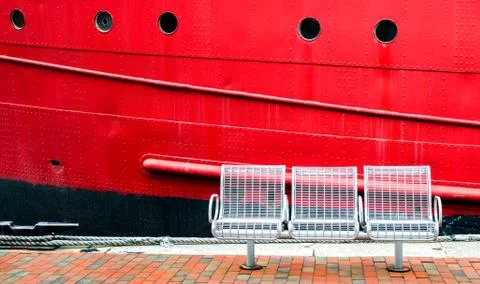 3 empty white seats on a brick walkway next to the hull of a red ship Stock Photos