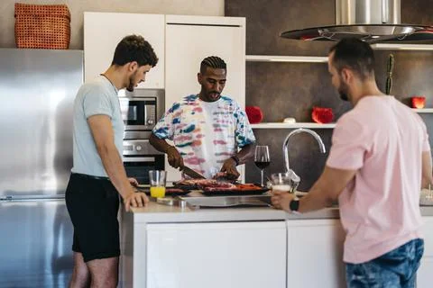 3 friends converse in a kitchen while one cuts meat with a knife Stock Photos