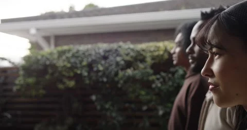 3 friends standing inline by yard hedge in hoodies, camera pulling back Stock Footage 344463200