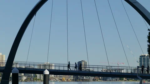 3 people cross a    single span pedestrian bridge - tall buildings in background Stock Footage 204054560
