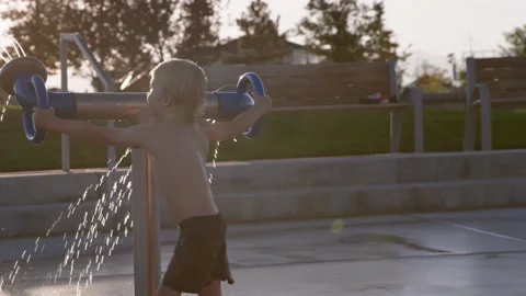 3 year old boy joyfully spins around with large splash pad water feature, sunset Stock Footage 233946208