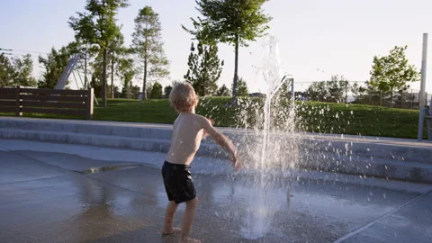 3 year old boy slaps water spraying upwards on splash pad at sunset, slow motion 스톡 동영상 233941231