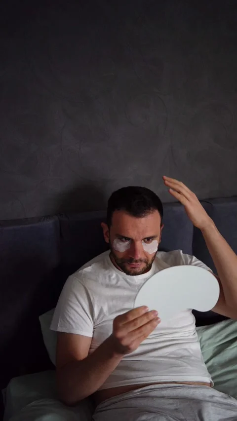 30-year-old man with a silk mask for sleep applying patches under the eyes .. Stock Footage 294524597