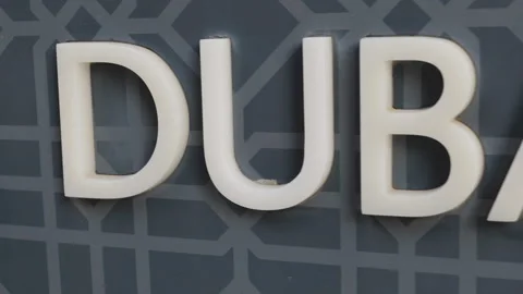 A 3D "DUBAI" sign mounted on a dark, geometrically patterned wall. Stock Footage 333472682