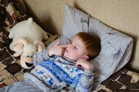 A 4-year-old red-haired boy is lying on the bed and picking his nose Stock Photos