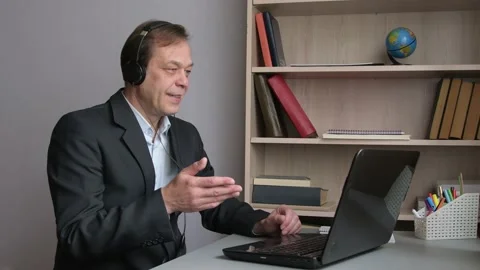  A 45-50-year-old man is sitting in the office at a laptop with headphones, comm Stock Footage 219333180