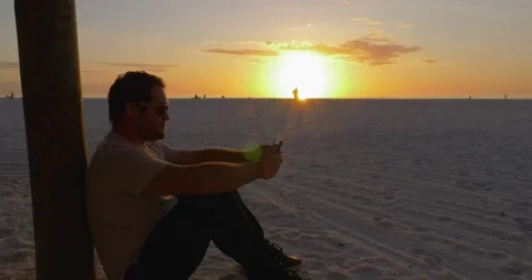 487 Man sitting on a beach during a sunset while on the phone Video stock 134546321