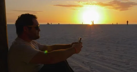 488 Closeup of a Man Sitting on a beach during a sunset while on the phone Stock Footage 134546633