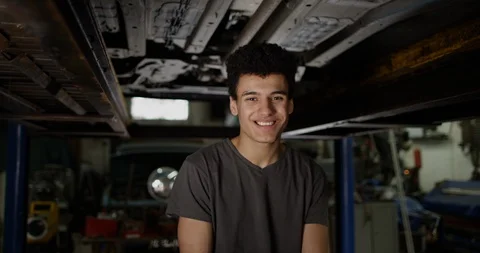 4K Apprentice garage mechanic standing under vehicle ramp &amp; smiling at camera Stock Footage 112960301