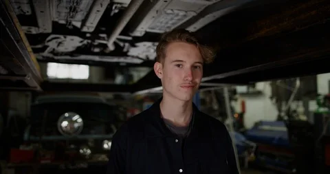 4K Apprentice garage mechanic standing under vehicle ramp &amp; smiling at camera Stock Footage 112968139