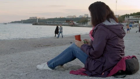 4K the beautiful lady in spring coat sits on the beach with cup of hot drink and Vidéo 100303821