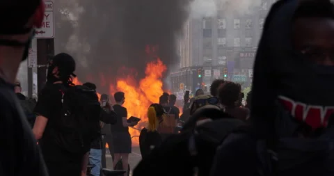 4K CAR ON FIRE WITH PROTESTERS IN FOREGROUND.  EXPLOSION GOES OFF IN DISTANCE Stock Footage 131453127
