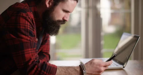 4K Casual man at home looking at smartphone with laptop open in front of him Stock Footage 61781710