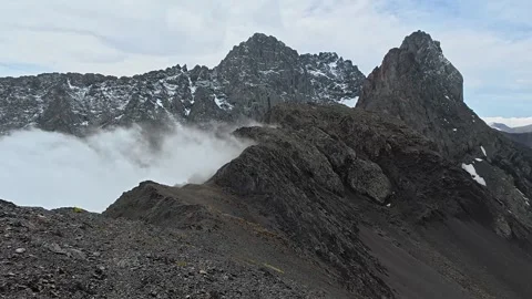 4K First-person view along a rocky ridge as clouds roll below the jagged, snow.. Видео 339462484