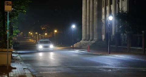 4K LONG SHOT OF EMPTY CITY STREET AT NIGHT WITH POLICE CAR DRIVING Video stock 131108575