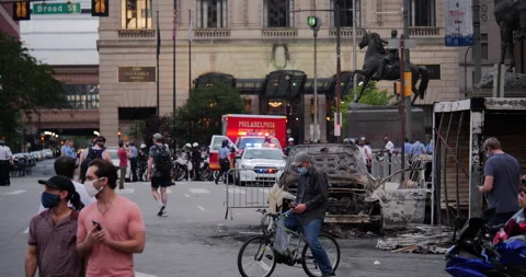 4K LONG SHOT OF PROTESTORS IN FACE MASKS PASSING BY BURNED OUT CAR Stock Footage 131459447