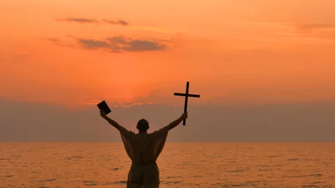 4К. Man ascetic, monk take cross  and Bible up  against sunset on seacost.  Stockbeeldmateriaal 102362913