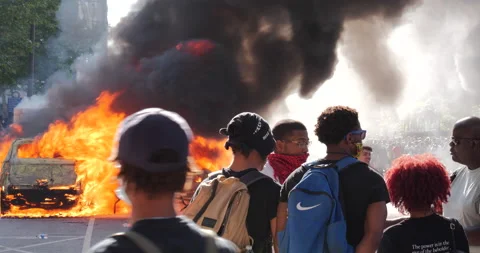 4K MEDIUM SHOT OF CAR ON FIRE WITH PROTESTERS TALKING IN THE FOREGROUND Stock Footage 131453775