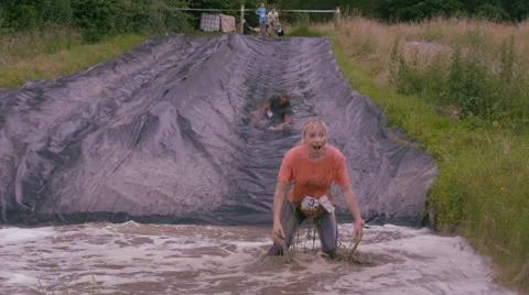 4K Mud race runners on assault course slide down hill into cold muddy water Stock Footage 66064322