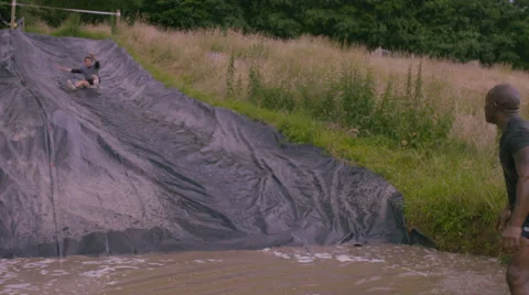 4K Mud race runners on assault course slide down hill into cold muddy water Stock Footage 66077541