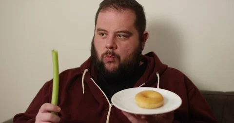 4K Overweight man on a diet making the choice to eat a celery stick or a donut Stock Footage