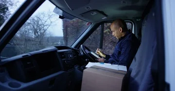 4K Portrait delivery driver smiling to camera, sitting in vehicle checking his p Stock Footage 85540779