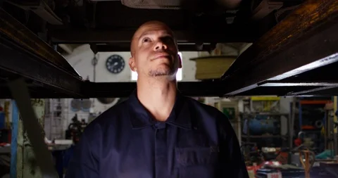 4K Portrait friendly smiling mechanic standing under vehicle in repairs shop Stock Footage 103827476
