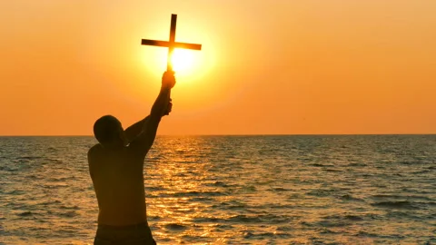 4К. Prayer man take cross up against marine orange sunset or sunrise .Back view Stockbeeldmateriaal 103676913