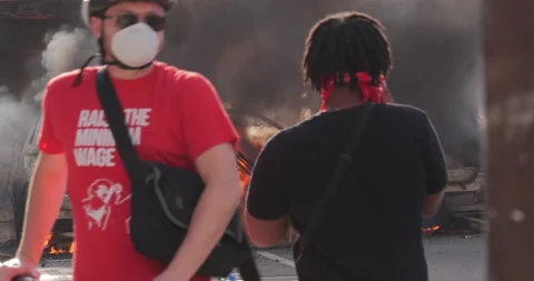 4K TWO MEN IN MASKS PASSING ONE ANOTHER IN FRONT OF BURNING CARS Stock Footage 131454771