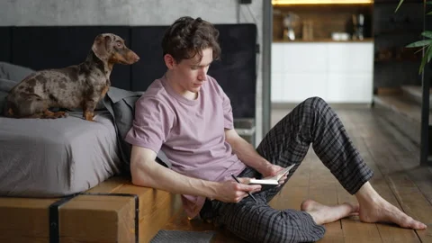 4k Young man is holding notebook and sitting on floor with dog in home interior Stock Footage 201396114
