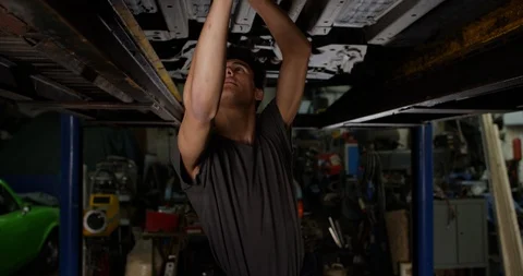 4K Young mixed race mechanic working on underside of car in garage workshop Stock Footage 112958155