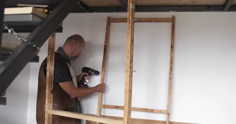 50 year old man assembling wooden shelf at home using battery powered drill Stock Footage 321880561